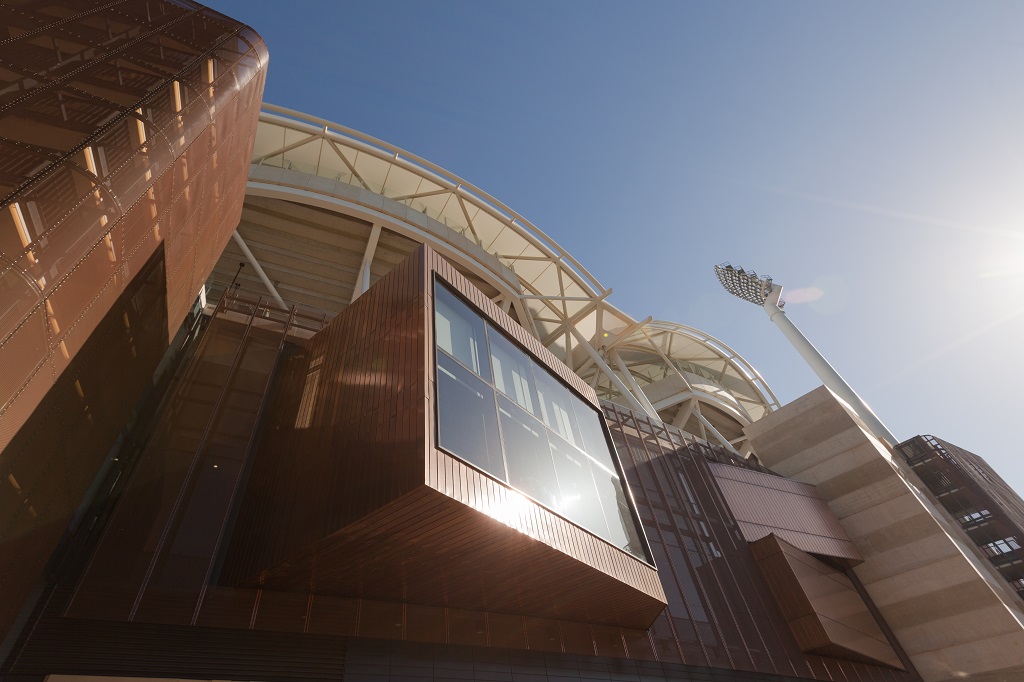 Exterior of the Adelaide Oval building. Geometric window extruded from the wall of building is covered with Nordic Bronze clad volumes. Walls are decorated with see through copper mesh. Sun is gleaming on the window glass and Nordic Bronze surface.