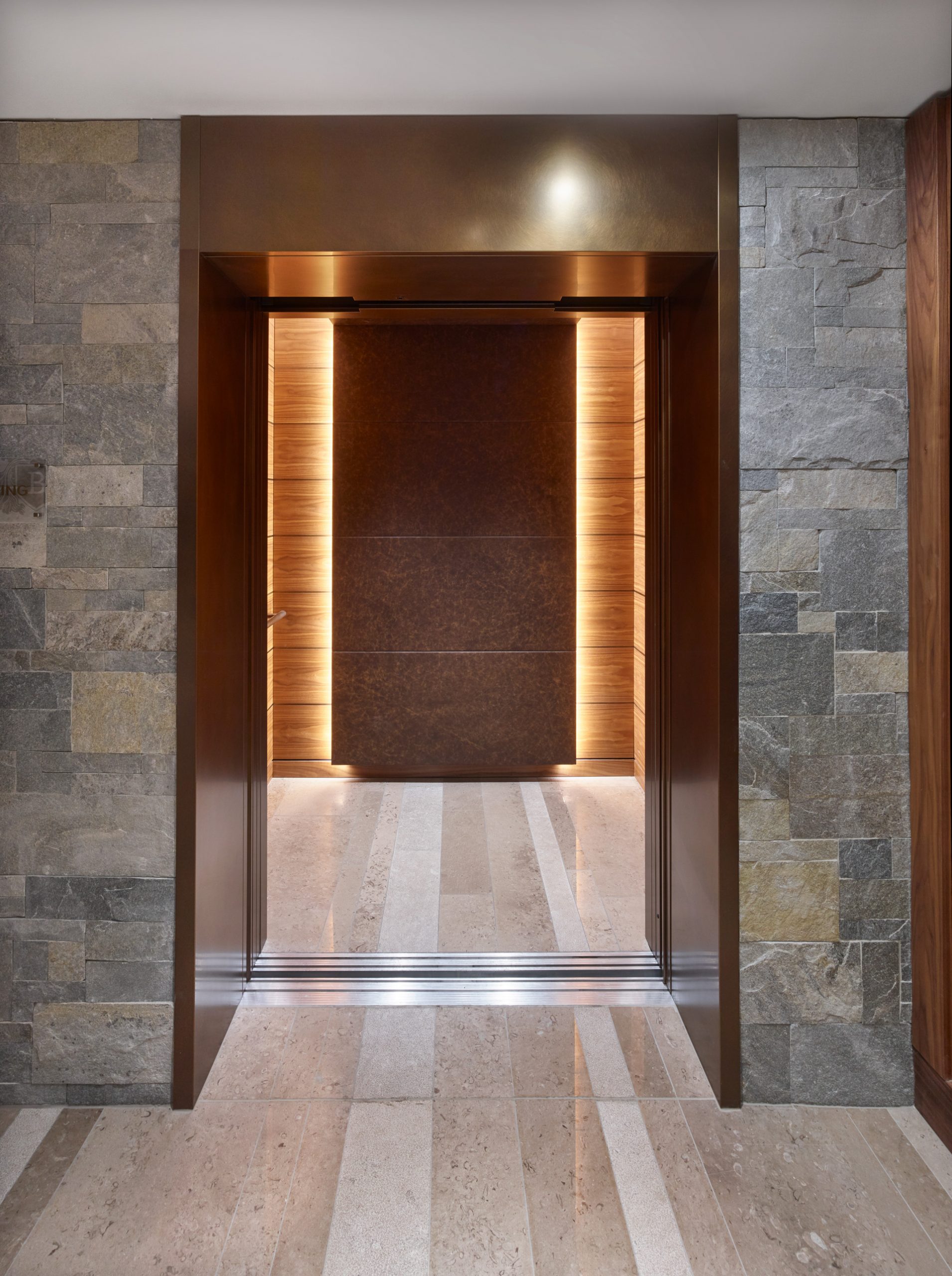 Doorway inside the Hotel Bürgenstock. The Doorway has semi-matter Nordic Décor copper finish while walls around it are stonework. Floor has reflective stone texture. The doorways leads to another corridor which has a copper element panel in the end of it. Copper element has a light source behind it.
