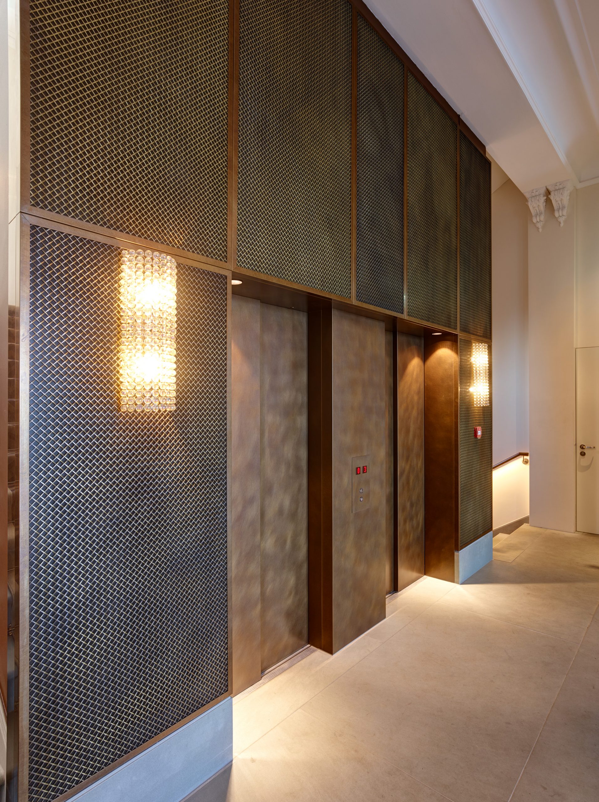 Elevators in a hall inside the Hotel Bürgenstock. Elevator doors are made from Nordic Décor copper and they have semi-matte surface. The walls of the elevator are also made from Nordic Décor. Top part of the elevator and walls on far right and far left have a copper mesh element on top of it. Walls on the far right and far left have decorated lamps mounted on their top parts.
