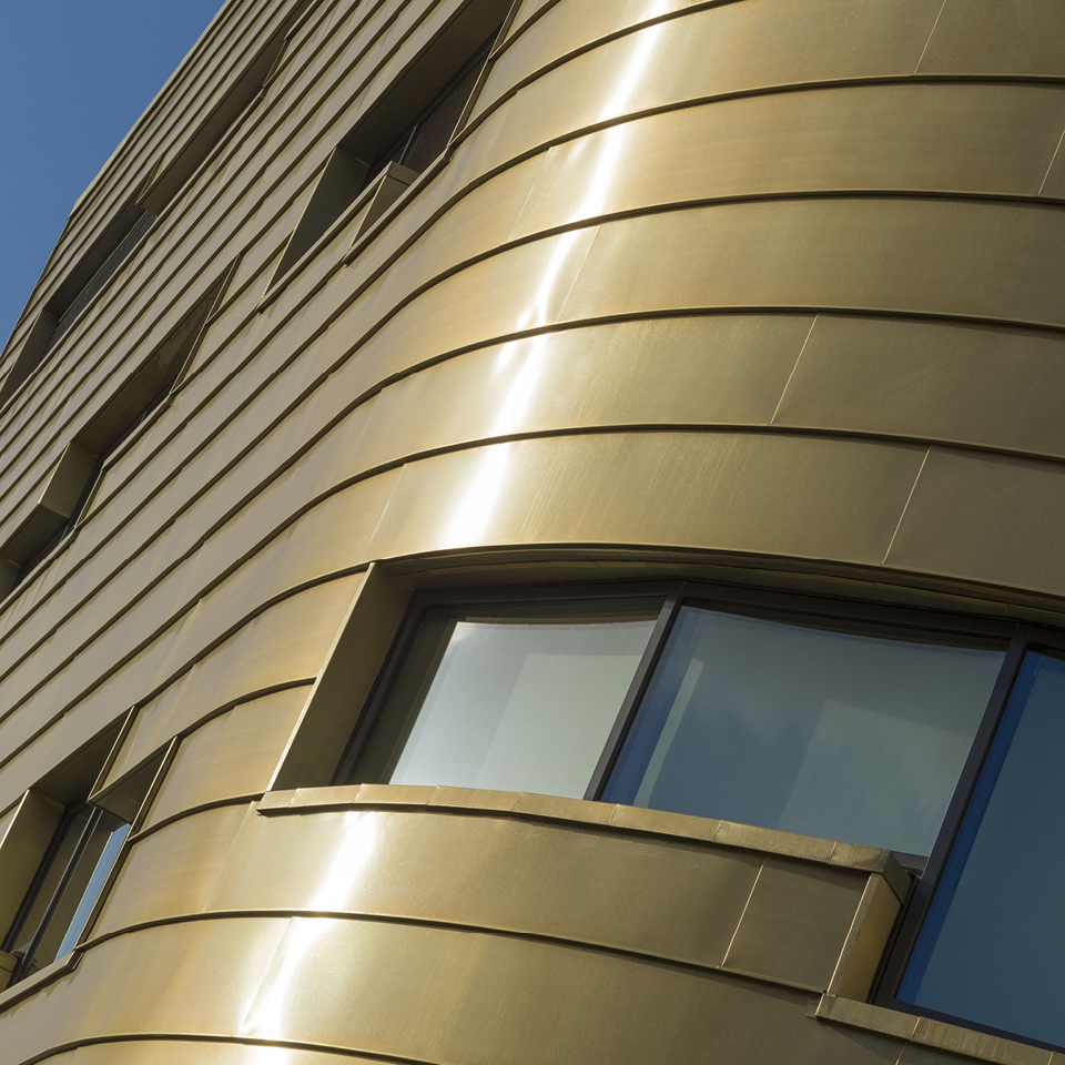 A close view of the curving copper wall of The Curve building located in Middlesbrough, UK. The wall is gold-coloured and its material is Nordic Royal Copper by Aurubis. There are multiple windows in the image. The copper panels are layered. Light is reflecting off the copper panels. A small amount of sky is visible in the top left corner of the image.