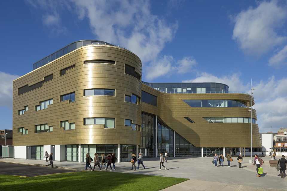 A view of the front yard of the Curve Building located in Middlesbrough, UK. The front side of the building is shown. The walls are clad from Nordic Royal Copper by Aurubis. The building has five floors. There are students walking in front of the building. It is daytime and the sky is cloudy. Light is reflecting off of the copper walls.