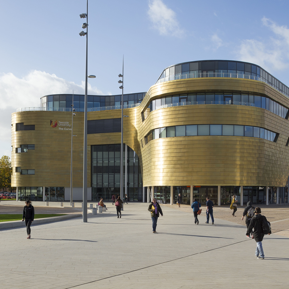 View from the yard of the Curve Building located in Middlesbrough, UK. The front side of the building is shown. The building walls are curving. Walls are layered and made from Nordic Royal Copper by Aurubis. People are walking in the front of the picture. Light is reflecting off the gold-coloured walls.