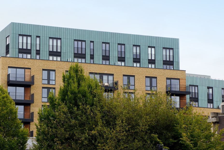 Exterior view of the Ram Quarter located in London, UK from a distance. Some trees in the forefront of the picture. Background has a large building with brick walls and copper walls. Copper walls are clad from Nordic Green Living 1 copper by Nordic Copper and Aurubis. Building has multiple floors and many windows.