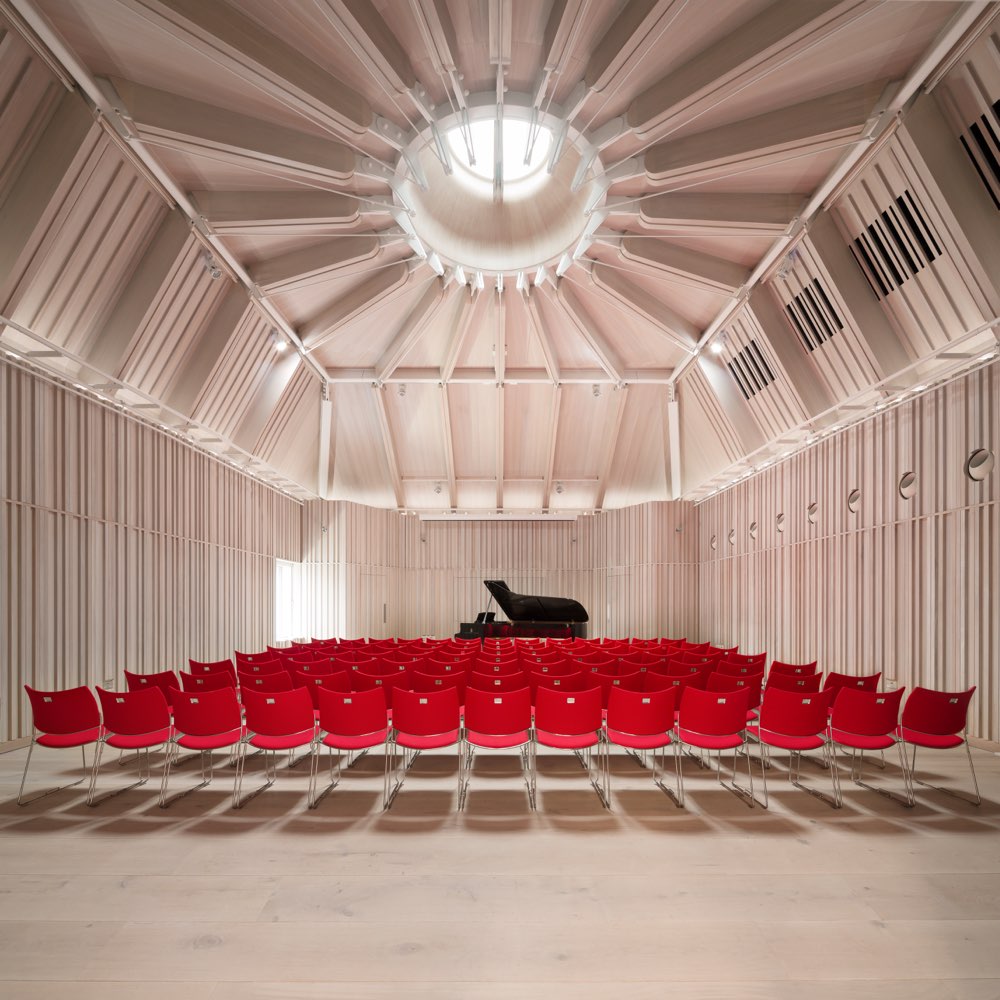 The interior of Royal Academy of Music located in London, UK. The room is a recital hall. The room has a black grand piano and a hundred red chairs facing the piano. The walls of the room are light brown coloured and have horizontal accents. The roof cascades upwards and has a spherical window at the top. The roof is made form Nordic Blue Copper by Aurubis.