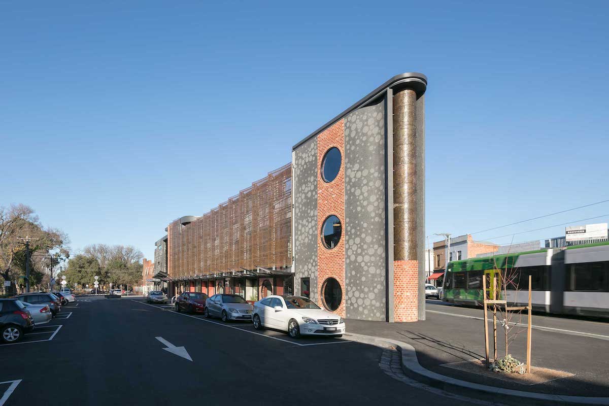 End part of the front facade of the North Fitzroy Library and Community Hub. Parking lot can be seen in front of the building and cars are parked there. In the end of the building, there is a two part pillar like shape. Pillar's top part is made from perforated Nordic Brass and bottom part is brickwork. Facade is 