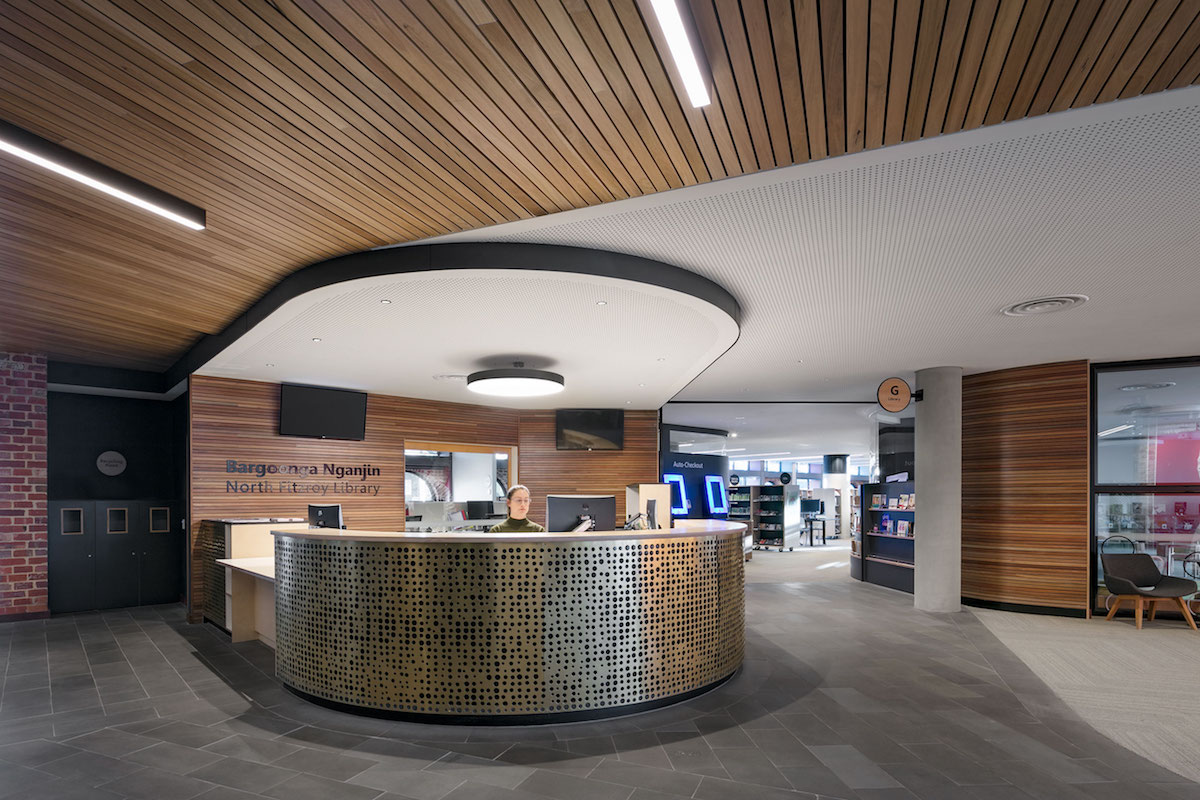 Lobby inside the North Fitzroy Library and Community Hub. Receptionist is seated behind a tall curved table which has perforated Nordic Brass paneling over it. Floor of the lobby is made from smoothed stone slabs. Walls are made from brickwork and wood paneling. Most of the forms in the overall architecture are curved.
