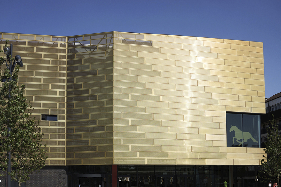 Wall and a window of the Deptford Lounge located in London, UK. Walls are clad from Nordic Royal Copper by Aurubis and Nordic Copper. Copper walls are gold-coloured and sunlight is reflecting from them.